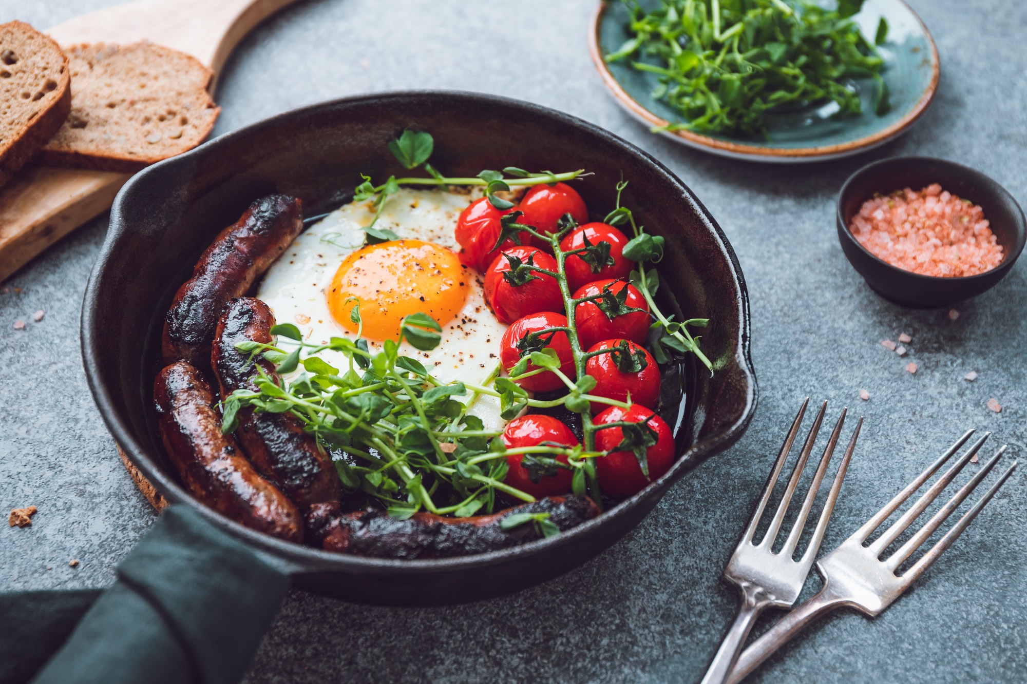 Breakfast time, fried egg with sausages and cherry tomatoes in a black iron pan, served microgreens.